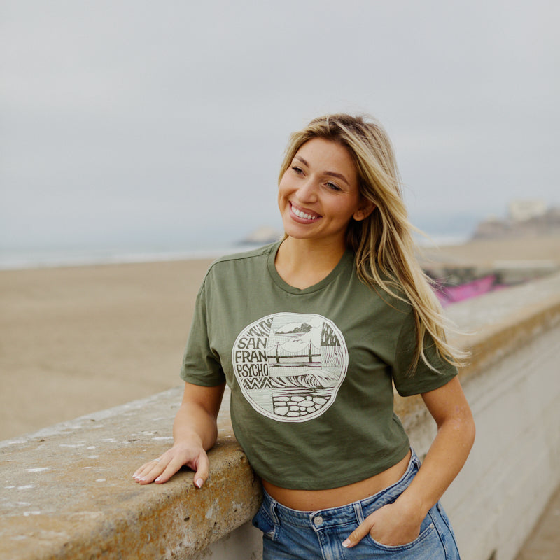 A woman smiles as she leans on a concrete railing at the beach wearing the Women's Elemental Cropped Tee, featuring a green fabric with a San Francisco-inspired graphic design.