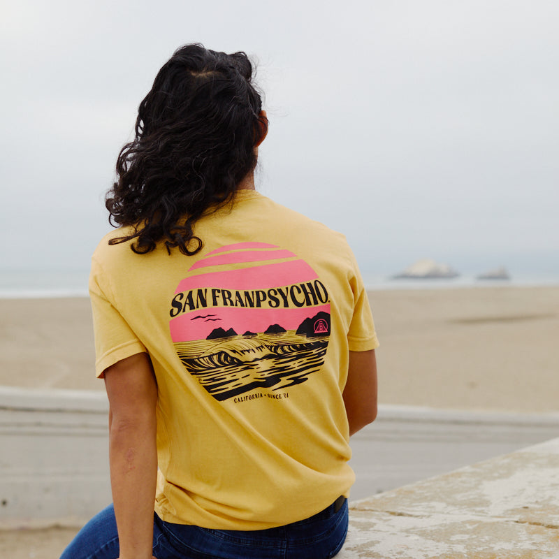 A person with long dark hair sits on a beach ledge, wearing the Kelly's Cove Tee, giving off timeless coastal city vibes.