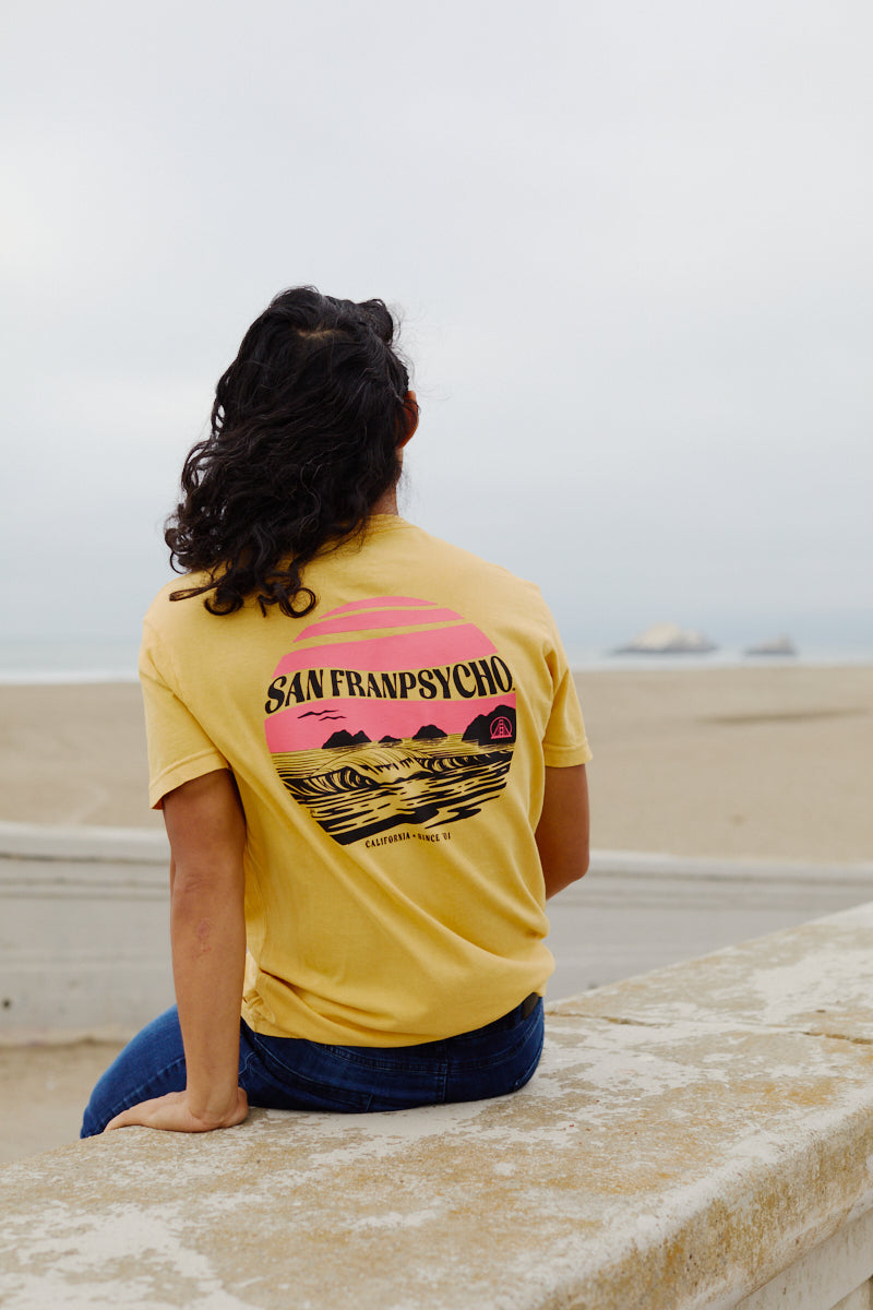 A person with long dark hair sits on a beach ledge, wearing the Kelly's Cove Tee, giving off timeless coastal city vibes.