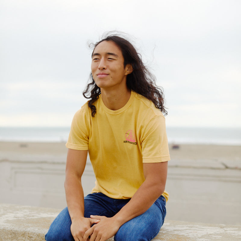 Wearing a yellow Kelly's Cove Tee, a person with long hair sits on a beachside ledge, smiling softly and enjoying the coastal city atmosphere.