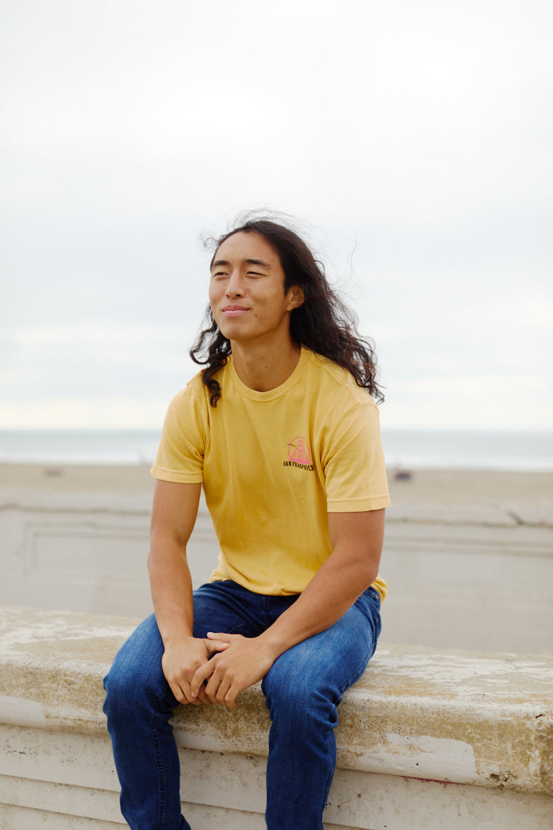 Wearing a yellow Kelly's Cove Tee, a person with long hair sits on a beachside ledge, smiling softly and enjoying the coastal city atmosphere.