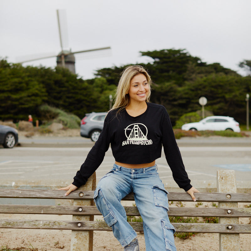 A smiling woman sits on a park bench in front of a windmill, radiating coastal vibes in the Women's Gull Logo Cropped Long Sleeve and cargo jeans.
