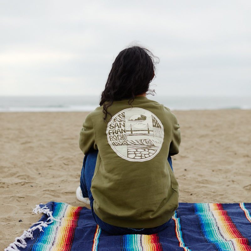 A person sits on a colorful blanket at the beach, facing the ocean, wearing a green Elemental Crewneck made from 100% cotton.