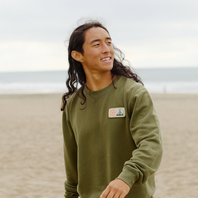 Wearing the Elemental Crewneck in green, a person smiles while walking along a sandy beach with the ocean behind them.