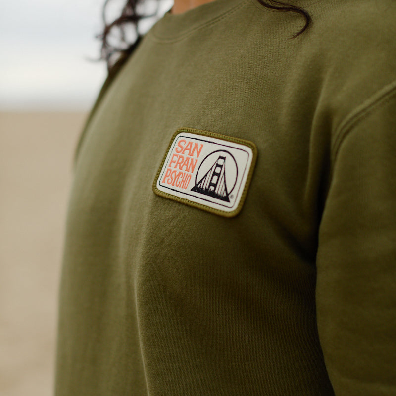 Person wearing the Elemental Crewneck in olive green, featuring a bridge illustration embroidered patch and made from 100% cotton.