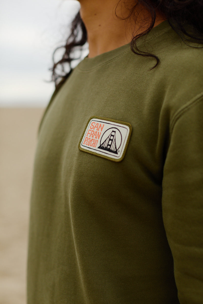 Person wearing the Elemental Crewneck in olive green, featuring a bridge illustration embroidered patch and made from 100% cotton.