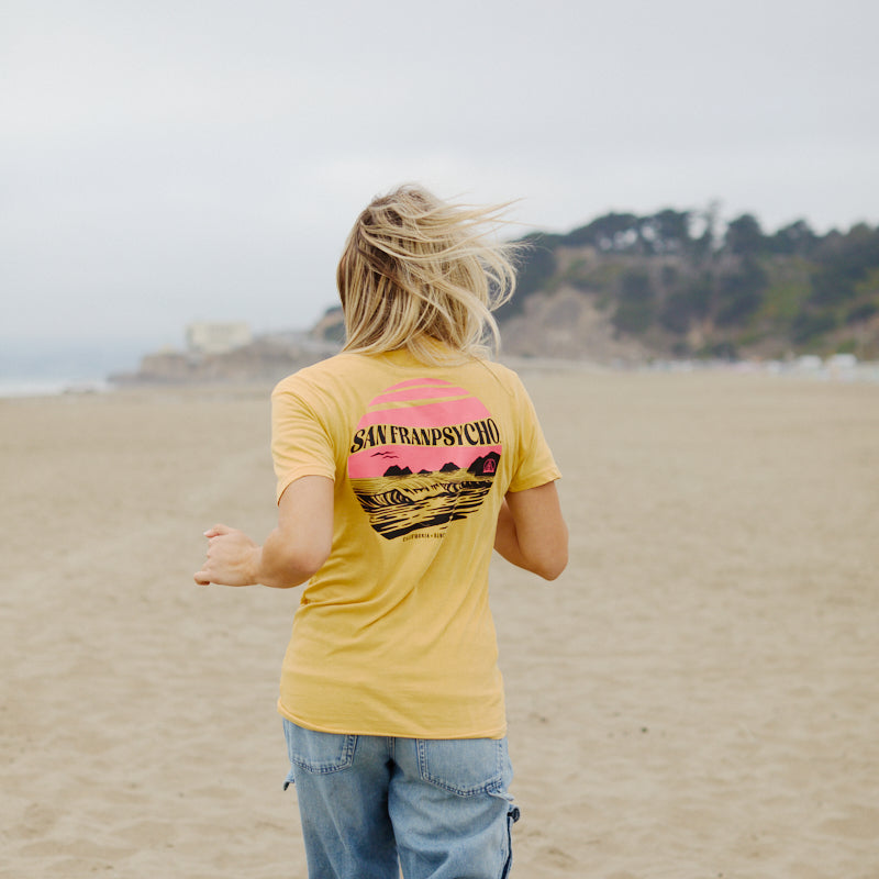 Person with long hair in a Kelly's Cove Tee walks on a sandy beach, facing away—capturing the true vibes of a coastal city.
