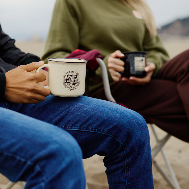 Two people relaxing on a sandy West Coast beach, sitting on outdoor chairs and holding Bear Essentials Mugs.