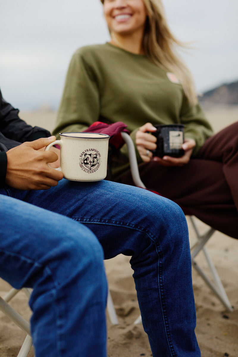 Two people relaxing on a sandy West Coast beach, sitting on outdoor chairs and holding Bear Essentials Mugs.