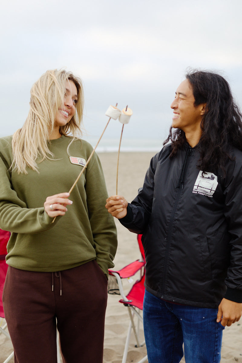 Two people smile and roast marshmallows on the beach in San Francisco, seated on chairs in the sand, both wearing cozy, wind-resistant Elemental Jackets.