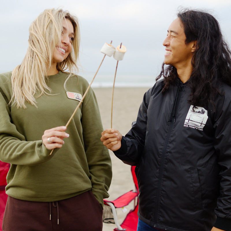 Two people in Elemental Crewnecks smile and toast marshmallows on sticks at a beach, with chairs and sand in the background.