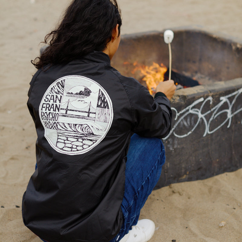 A person roasts a marshmallow over a beach fire pit, staying cozy in the wind-resistant Elemental Jacket featuring a San Francisco graphic.