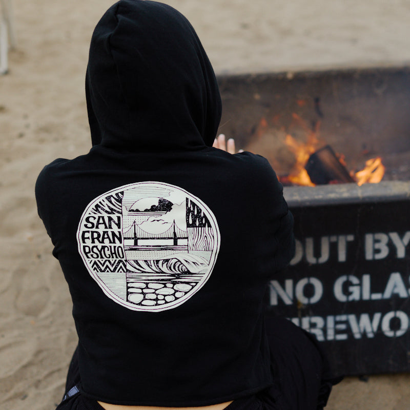 Someone in an Elemental Crop Hoodie featuring a circular San Franpsycho design sits by a beach fire pit.