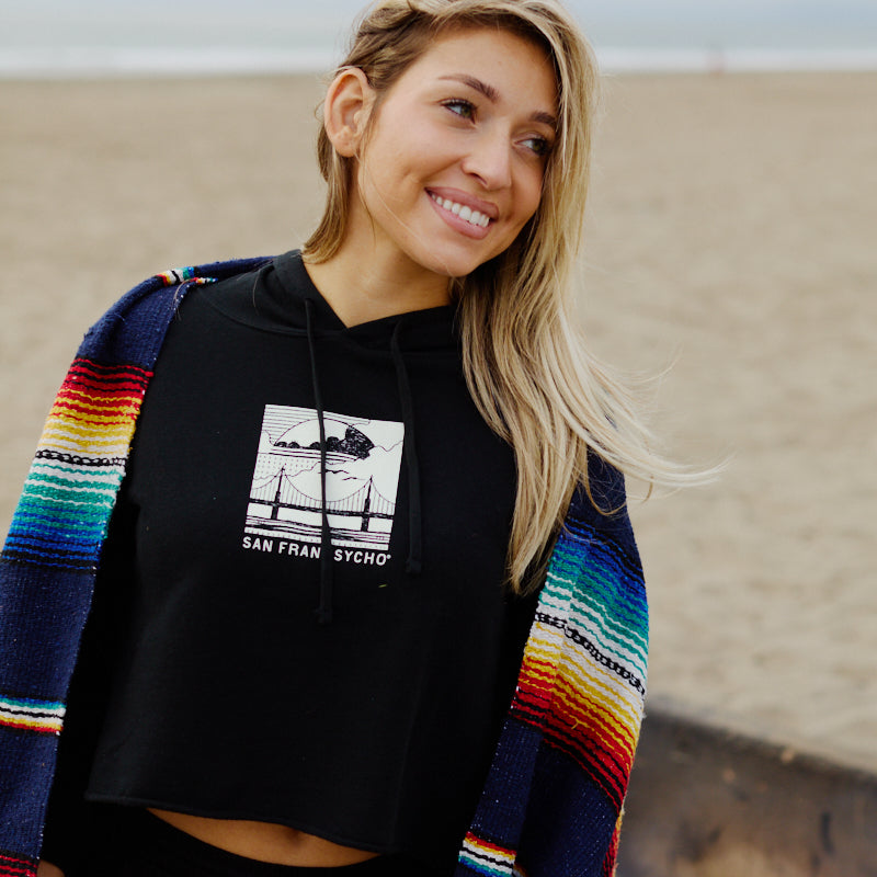 A smiling woman wearing the Elemental Crop Hoodie and holding a colorful blanket stands by a fire pit on a sandy beach.