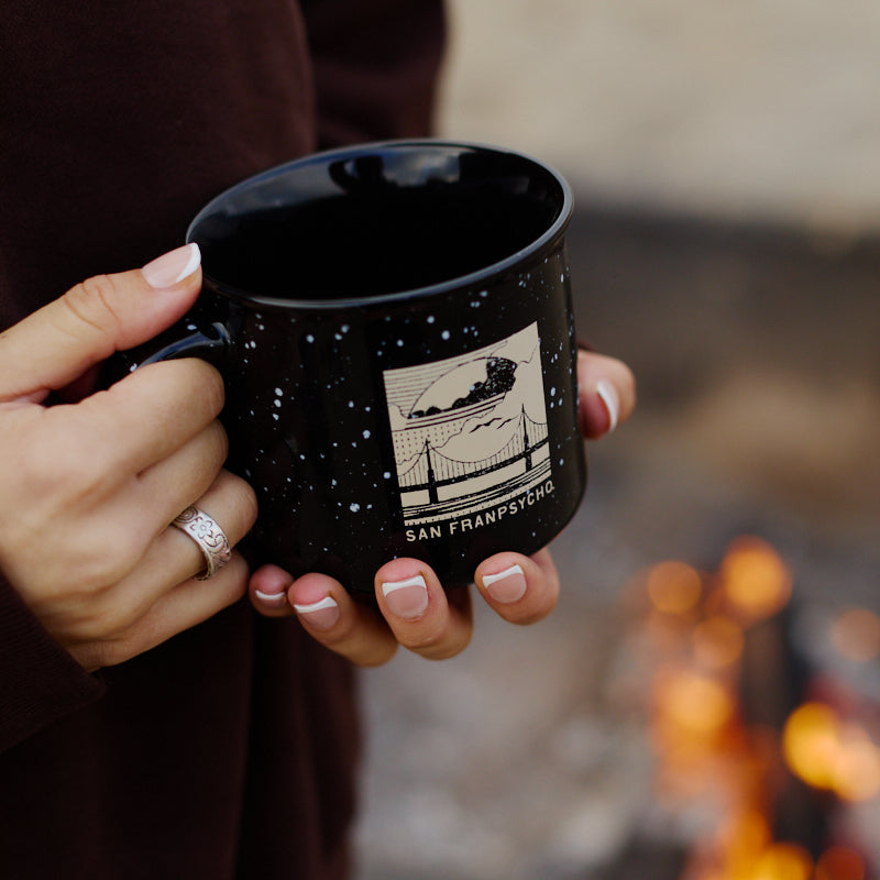 A person holds an Elemental Mug—a black campfire mug featuring a San Francisco design—near a fire pit while wearing a dark sweater.