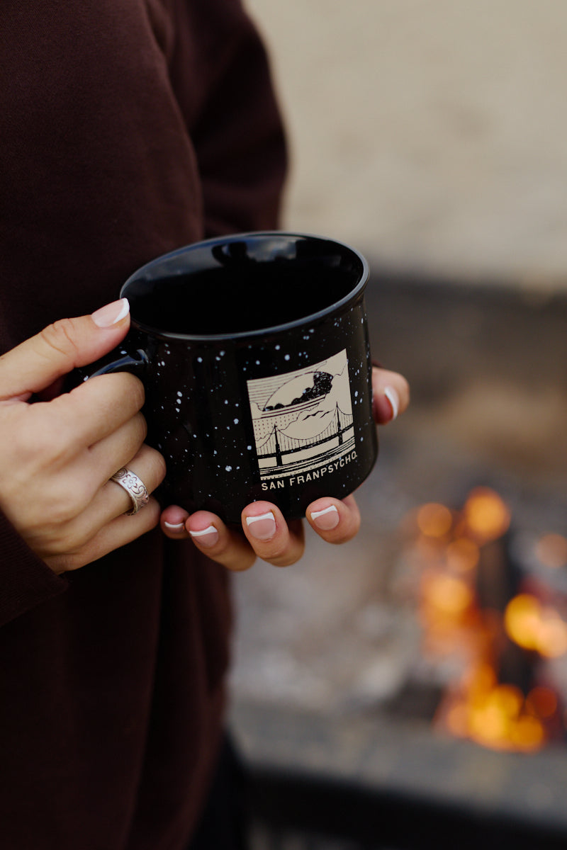 A person holds an Elemental Mug—a black campfire mug featuring a San Francisco design—near a fire pit while wearing a dark sweater.