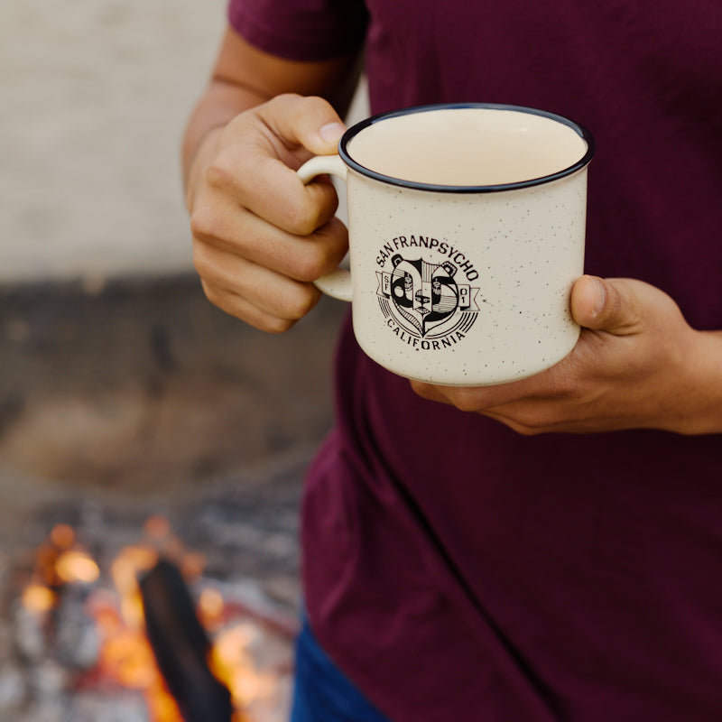 A person in a maroon shirt holds the Bear Essentials Mug—a speckled ceramic mug featuring San Francisco, California—by a campfire, capturing a cozy West Coast vibe.