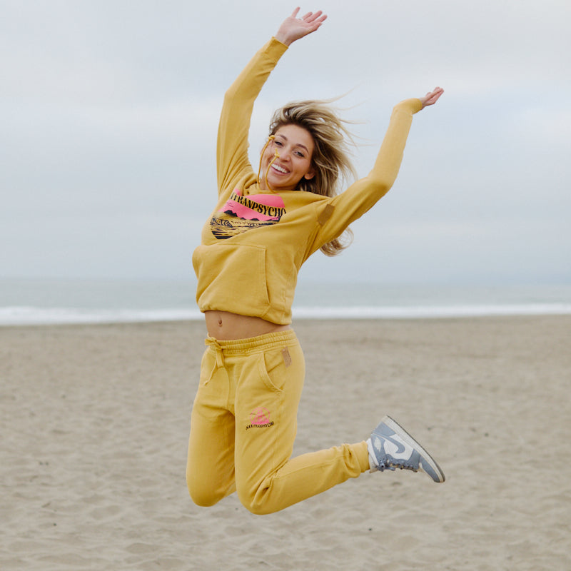 A woman in a Kelly's Cove Pullover Hoodie jumps joyfully on a sandy beach, capturing coastal city vibes with the ocean and cloudy sky in the background.