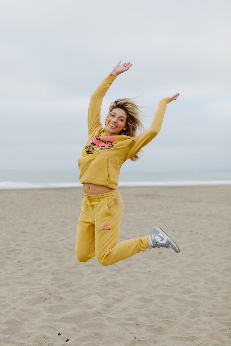 A woman in a Kelly's Cove Pullover Hoodie jumps joyfully on a sandy beach, capturing coastal city vibes with the ocean and cloudy sky in the background.