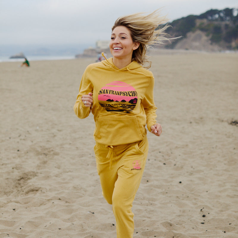 A woman jogs along the sandy beach, smiling with windblown hair and easy coastal vibes in a hand-printed Kelly's Cove Pullover Hoodie.