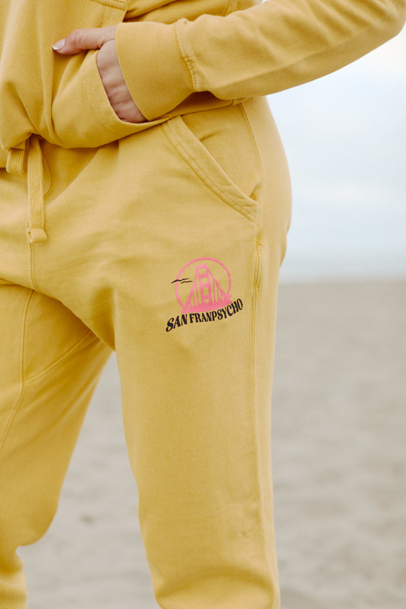 Someone wearing Kelly's Cove Joggers in yellow with a pink and black San Franpsycho logo, standing on the sandy beach.