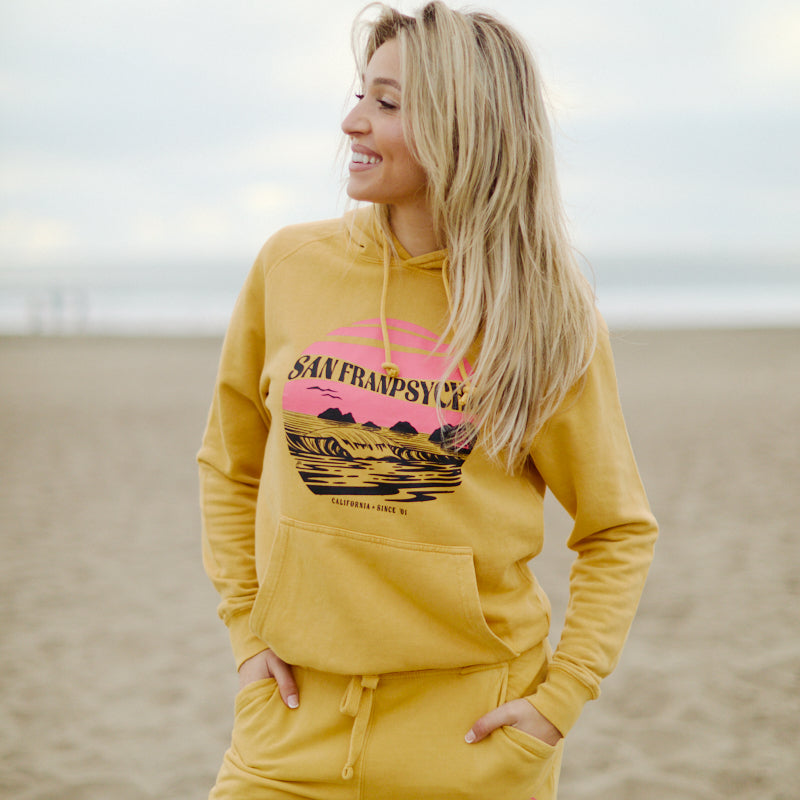 A woman in a hand-printed yellow Kelly's Cove Pullover Hoodie smiles while standing on a sandy beach, capturing coastal city vibes.
