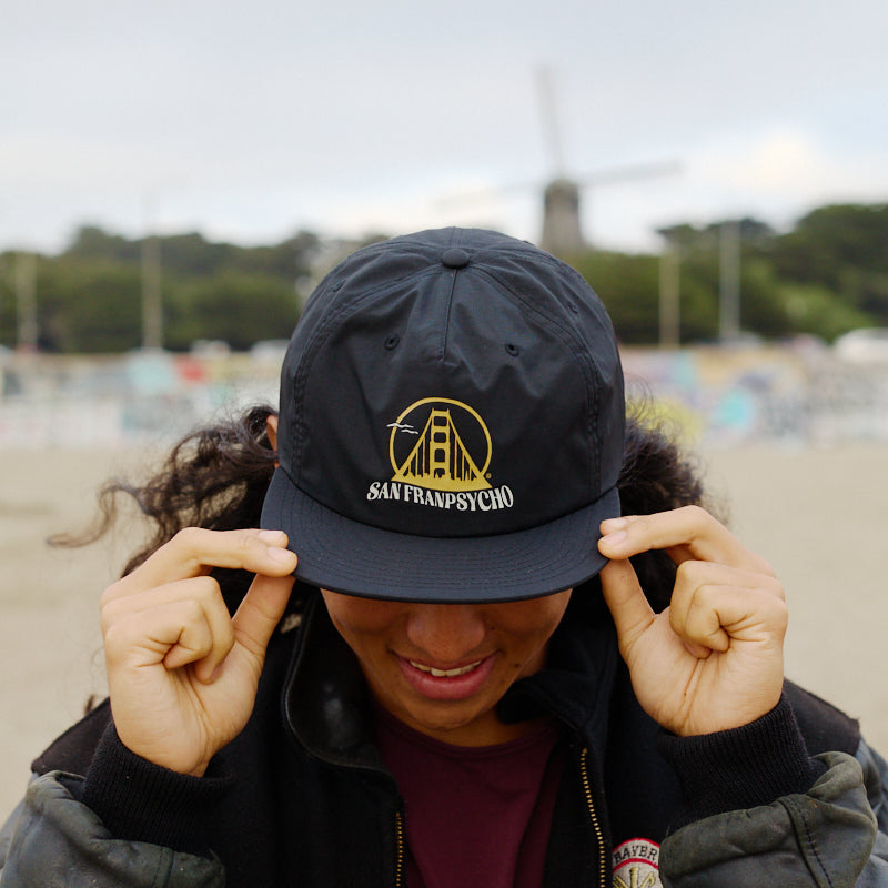 A person smiles and holds the flat brim of a Gull Logo Hat with an unstructured crown, as a windmill stands in the background.
