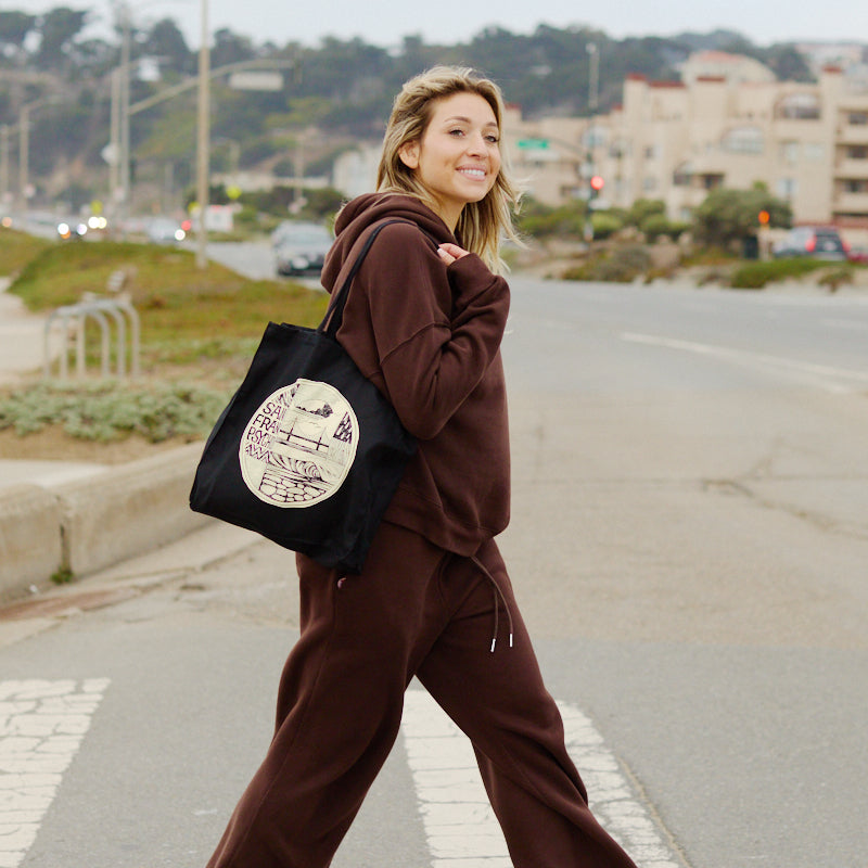 A woman in a brown tracksuit smiles while crossing a street, carrying the Elemental Tote featuring bold Erik Abel artwork.