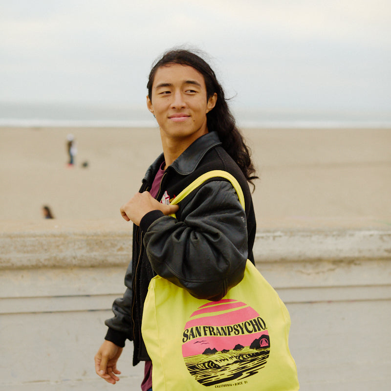 A person with long hair smiles at the camera on a San Francisco beach, holding a yellow Kelly's Cove Tote by San Franpsycho.
