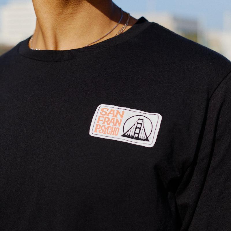 A person wearing a black Elemental Long Sleeve, hand printed by San Franpsycho, featuring a patch with the Golden Gate Bridge.
