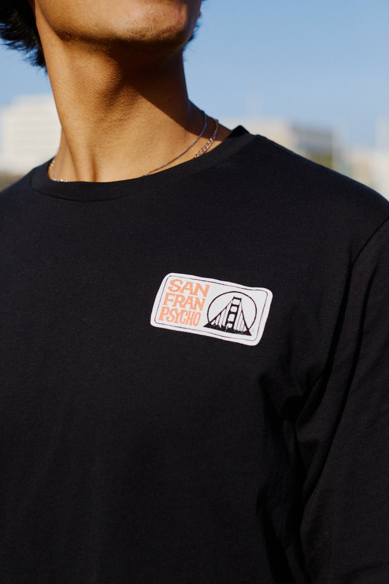 A person wearing a black Elemental Long Sleeve, hand printed by San Franpsycho, featuring a patch with the Golden Gate Bridge.