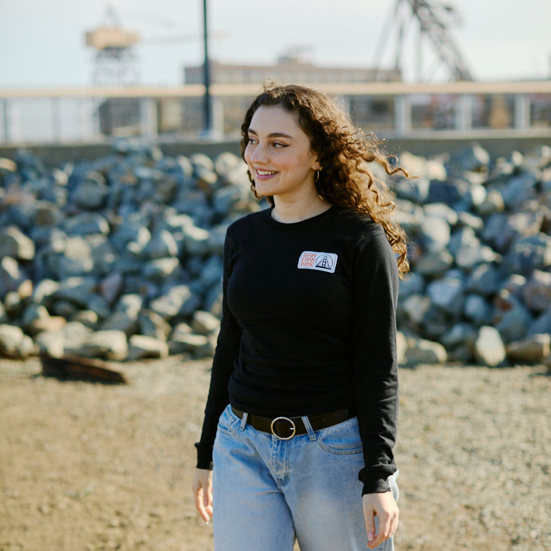 Smiling woman with curly hair in a black Elemental Long Sleeve and jeans stands outdoors near rocks on a sunny day.