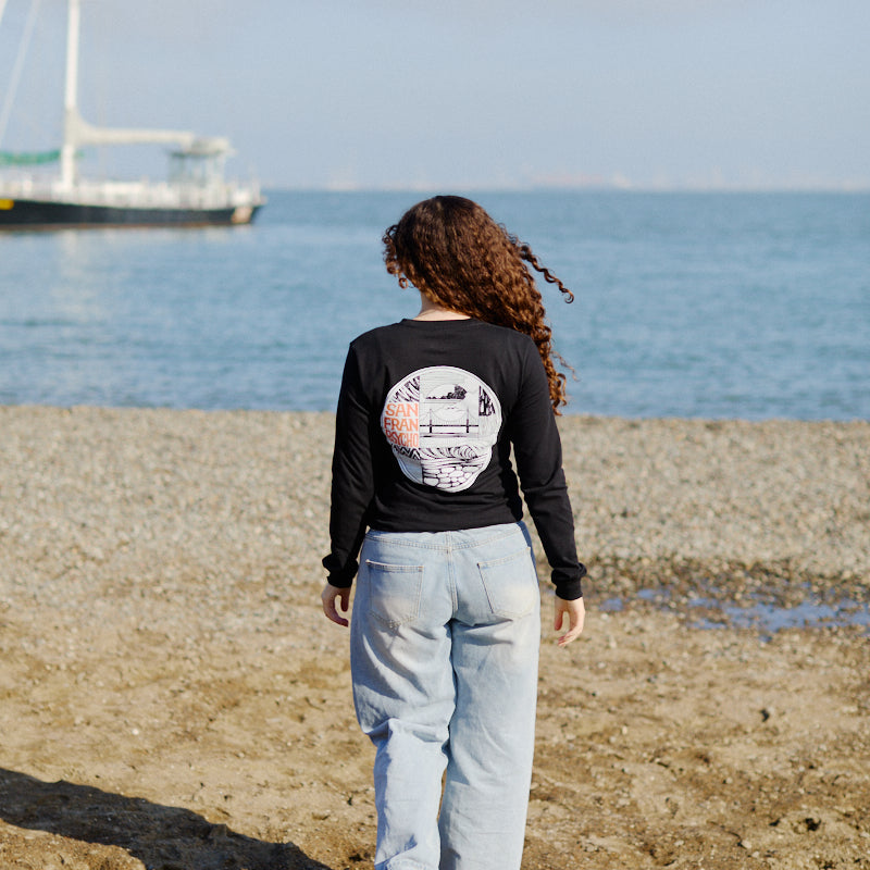 A woman with curly hair walks on a sandy beach toward the sea, dressed in jeans and the Elemental Long Sleeve.