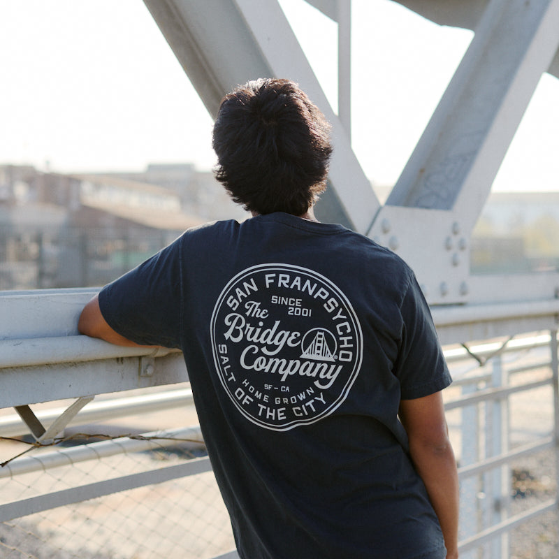 Wearing the Salt of the City Tee, hand printed in San Francisco, a person in jeans leans on a bridge railing, facing away from the camera.