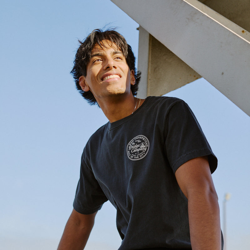 A young man in the Salt of the City Tee and jeans sits outdoors, smiling as he looks up under a metal structure.