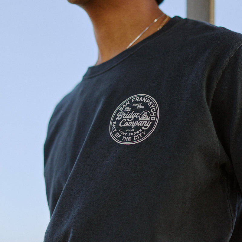 Smiling person wearing the Salt of the City Tee, a dark heavyweight cotton shirt with a round Straight Up Company logo hand-printed in San Francisco on the chest.