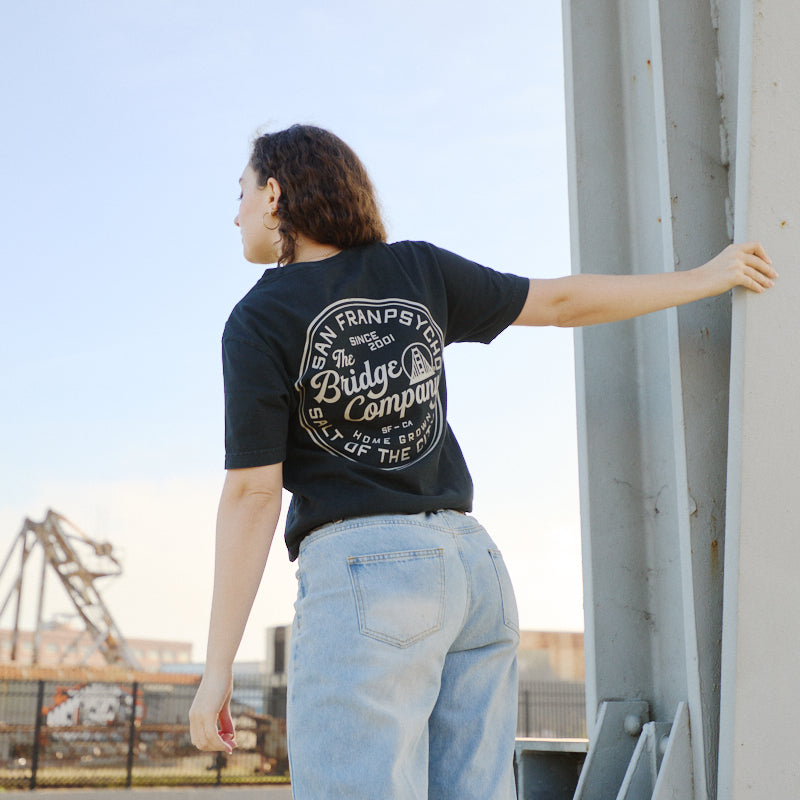 Wearing the Salt of the City Tee, hand printed in San Francisco, a person stands outdoors in jeans holding a metal beam near industrial structures.