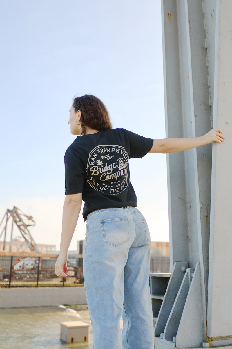 Wearing the Salt of the City Tee, hand printed in San Francisco, a person stands outdoors in jeans holding a metal beam near industrial structures.