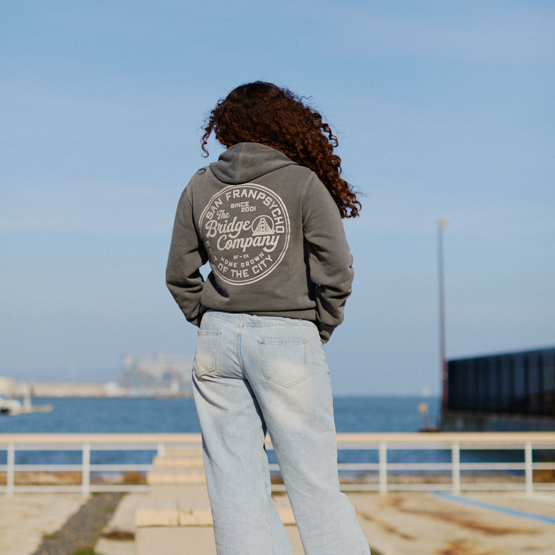 A person with curly hair in jeans and a Salt of the City Pullover Hoodie stands on dockside planks, gazing at the ocean under a clear sky.