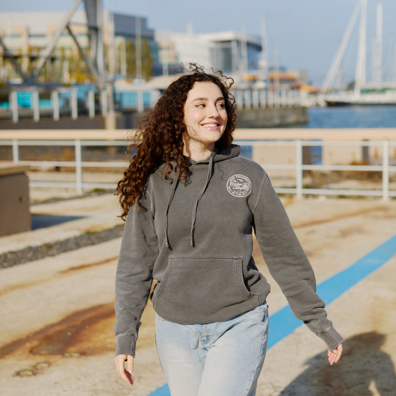 A smiling woman wears the Salt of the City Pullover Hoodie while walking outdoors near a waterfront on a sunny day.