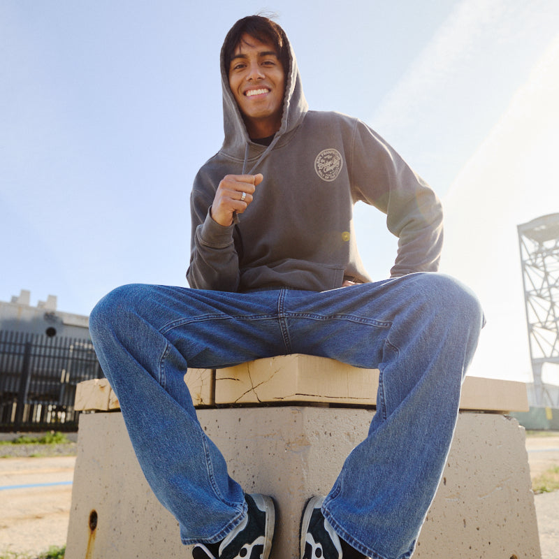 A smiling young man wearing the Salt of the City Pullover Hoodie and jeans sits on a concrete block outdoors, sunlight shining behind him.
