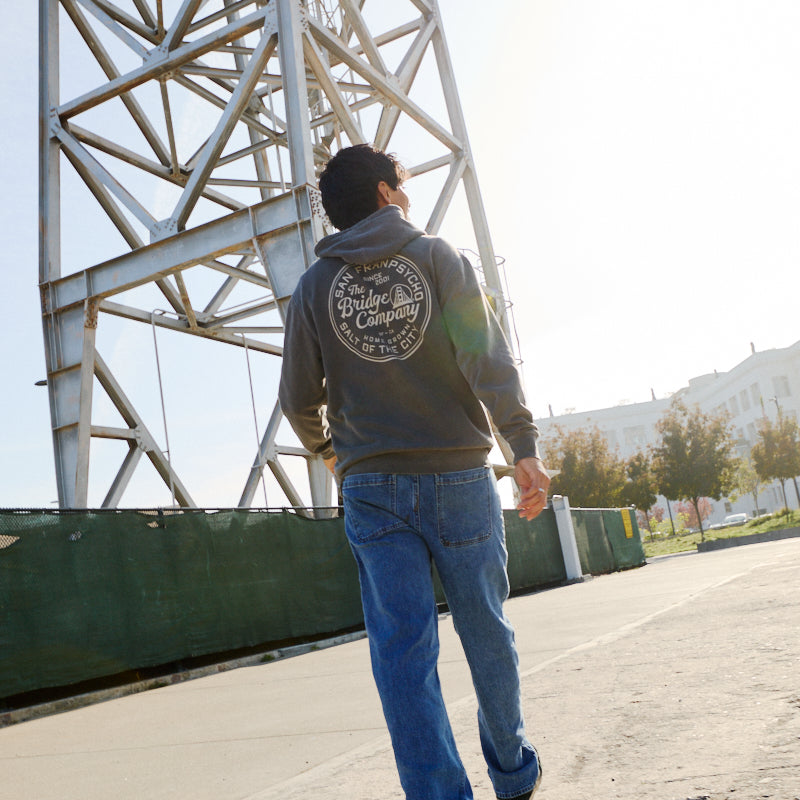 Wearing a Salt of the City Pullover Hoodie and jeans, a person strolls near a tall metal structure on a sunny day.