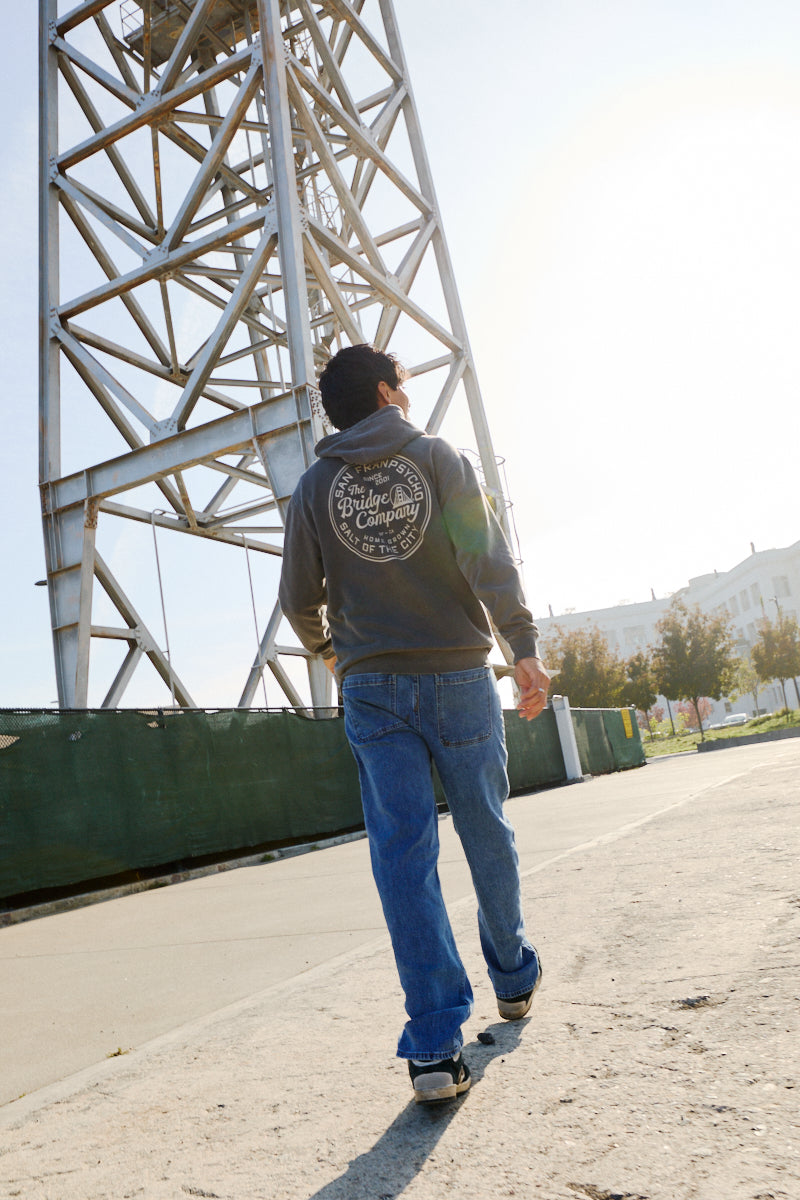 Wearing a Salt of the City Pullover Hoodie and jeans, a person strolls near a tall metal structure on a sunny day.
