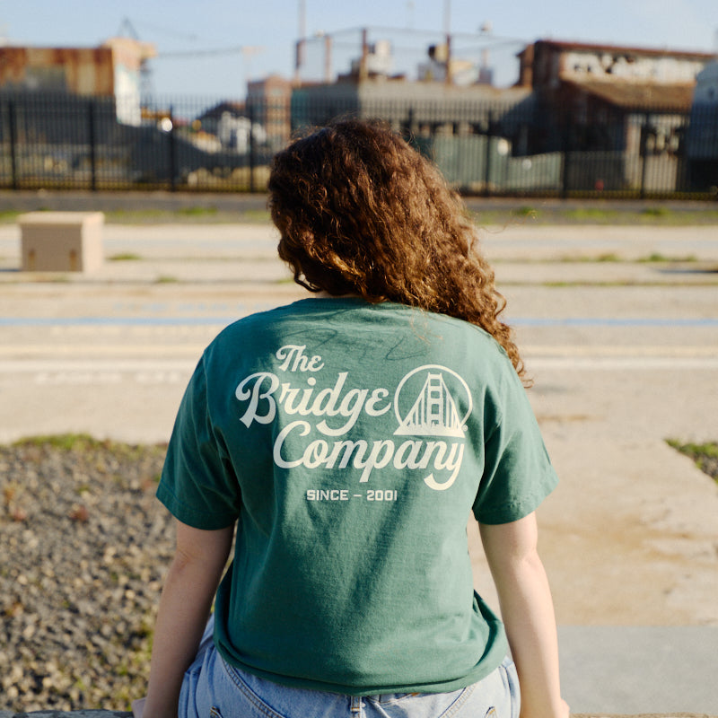A person with curly hair sits outdoors, wearing a green hand-printed Bridge Co Tee and denim shorts.