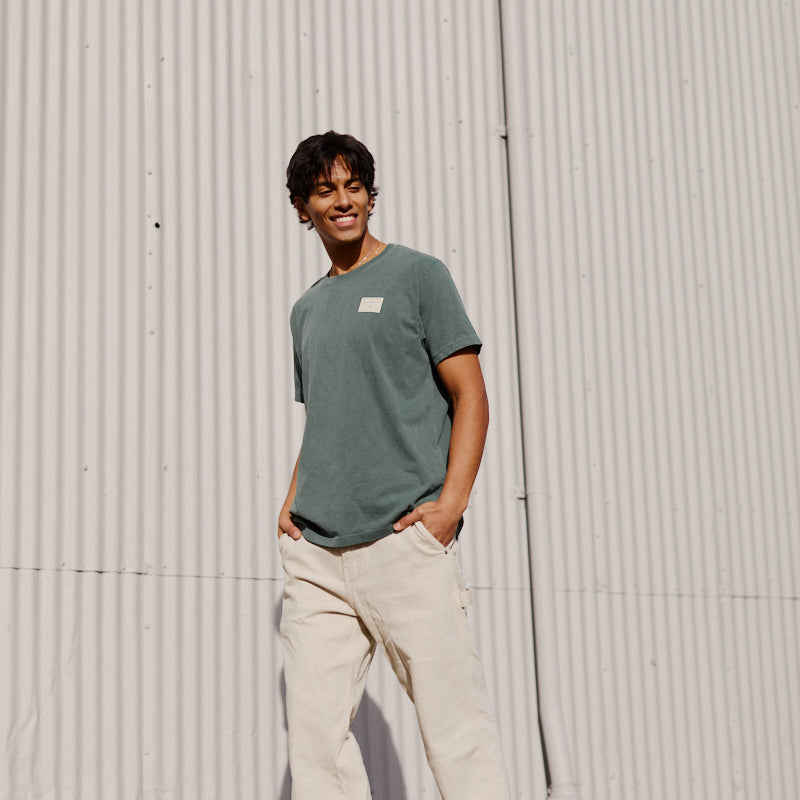 A person wearing the Bridge Co Tee in green stands outside against a corrugated metal wall, smiling.