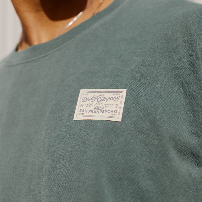 A person wears the Bridge Co Tee, a heavyweight green cotton shirt with a beige "The Bridge Company, San Franpsycho" patch label on the chest—ideal for workwear style.