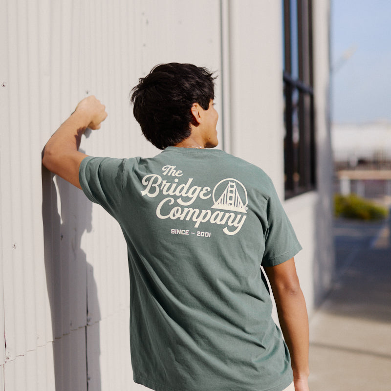 Wearing a Bridge Co Tee, a person stands outdoors facing a corrugated metal wall.