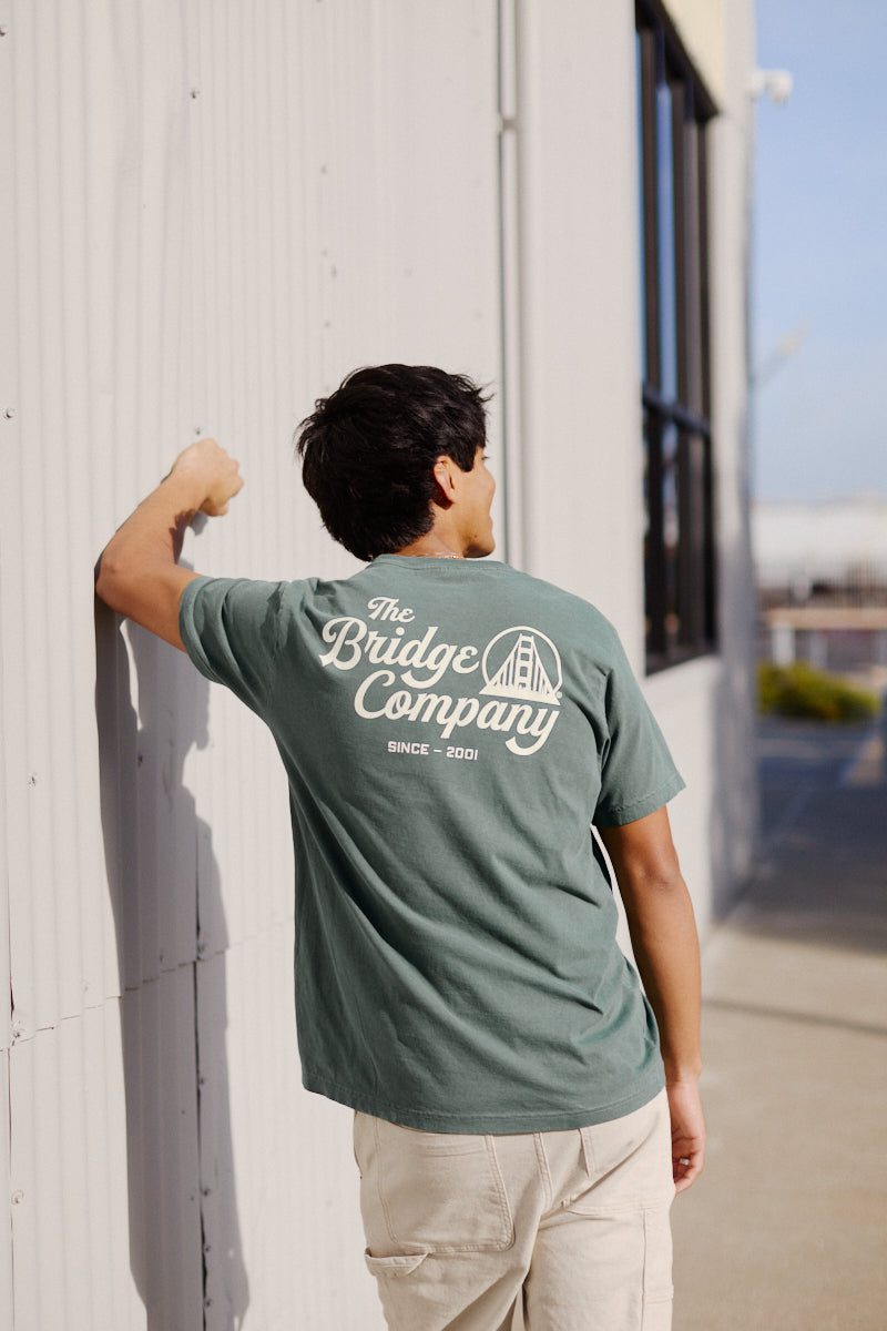 Wearing a Bridge Co Tee, a person stands outdoors facing a corrugated metal wall.