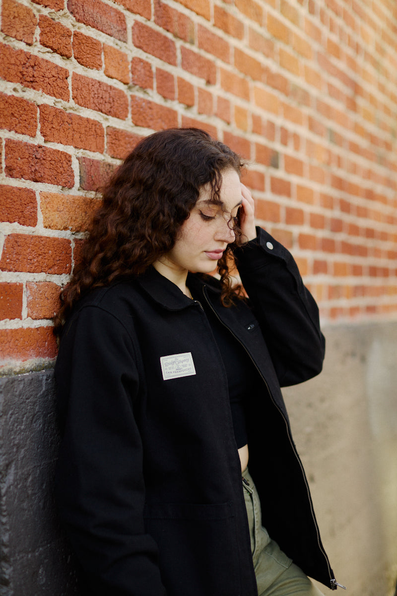A young woman with curly hair in the Bridge Co. Chore Jacket leans against a brick wall, looking down thoughtfully—embodying the timeless spirit of San Francisco-made workwear.