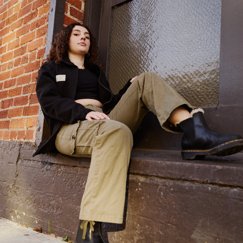 A person with curly hair sits on a stoop by a brick wall, wearing rugged workwear: boots, khaki pants, and the classic Bridge Co. Chore Jacket made in San Francisco.